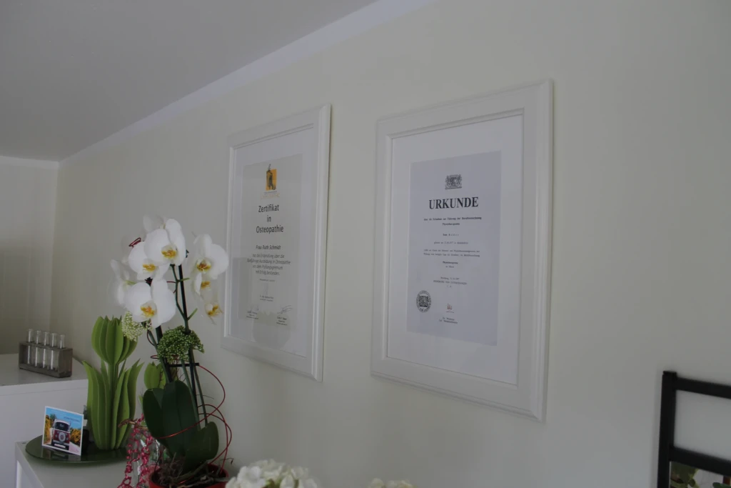 Weiße Wand mit gerahmtem Zertifikat in Osteopathie und Urkunde, davor weiße Orchidee